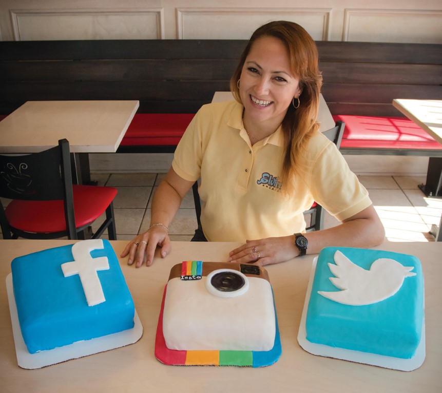 The owner of Elvira's seated in the bakery behind three custom cakes shaped like social media logos